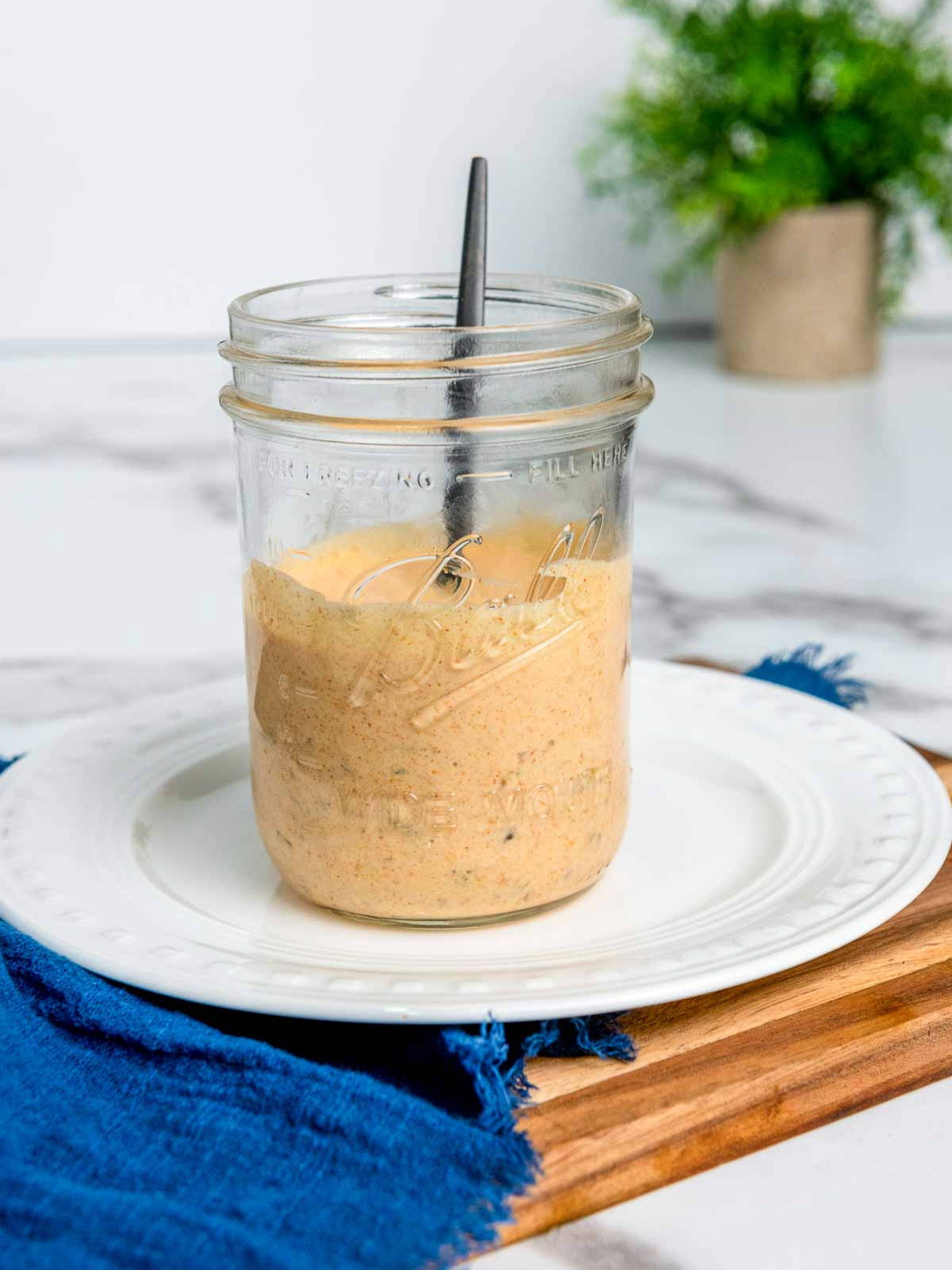 Glass mason jar filled with creamy orange burger sauce (Big Mac–style) with visible seasoning specks, a spoon inside, placed on a white plate over a wooden board with a blue cloth underneath.