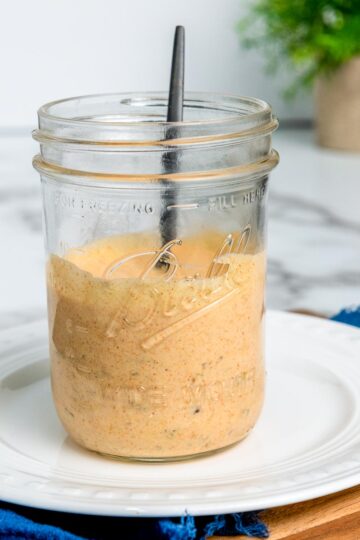 Homemade Big Mac copycat sauce in a glass mason jar with a spoon, placed on a white plate over a wooden board and blue cloth.