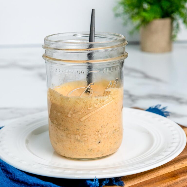 Homemade Big Mac copycat sauce in a glass mason jar with a spoon, placed on a white plate over a wooden board and blue cloth.