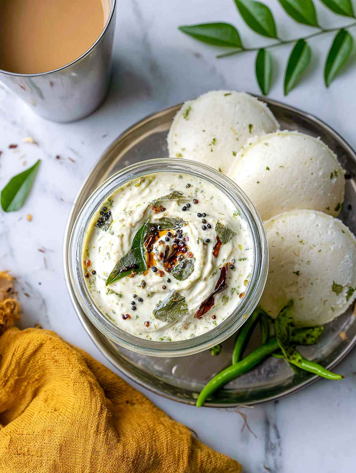 A glass jar of creamy coconut chutney topped with mustard seeds, curry leaves, and dried red chilies sits on a steel plate with soft idlis and green chilies, alongside a cup of chai.