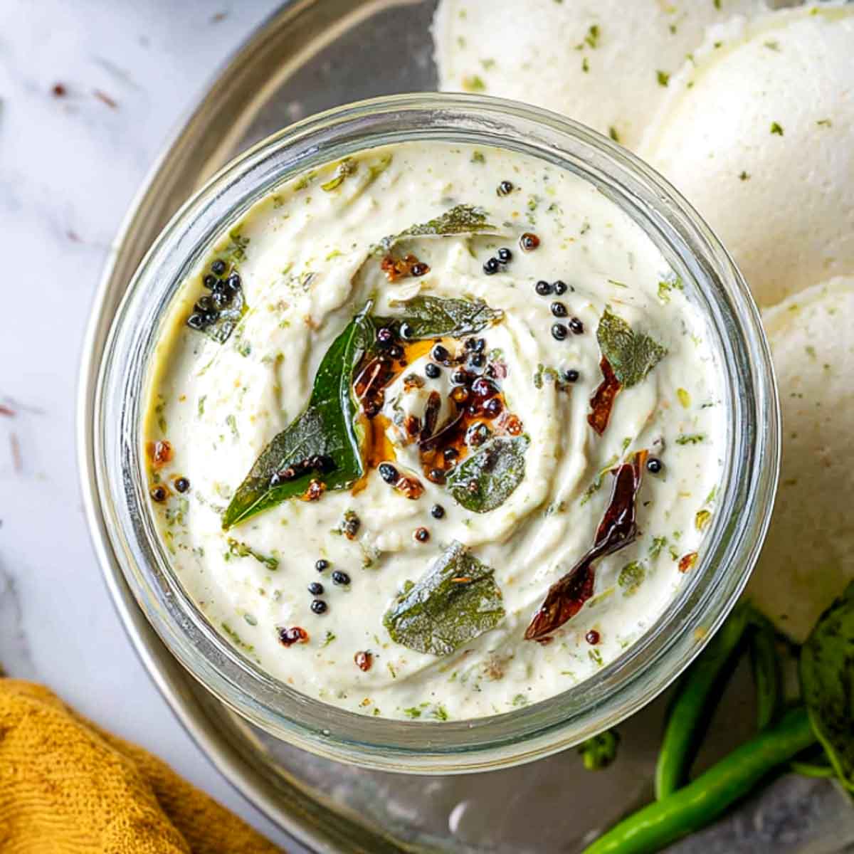 Close-up of smooth coconut chutney in a glass jar, finished with a tempering of black mustard seeds, curry leaves, and dried red chilies in hot oil.