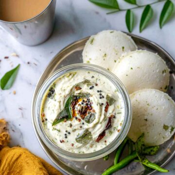 Overhead view of coconut chutney served in a glass jar with idlis on a steel plate, fresh curry leaves, green chilies, and a cup of tea on a marble surface.