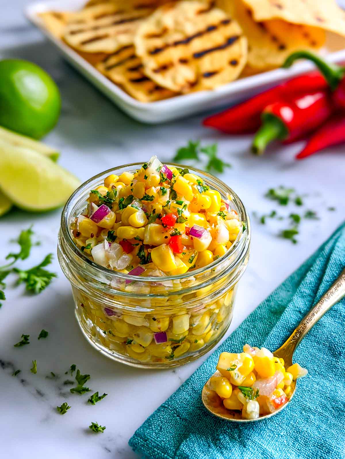 A small glass jar filled with bright homemade corn relish made with sweet corn, red onion, red pepper, and fresh herbs, set on a white marble surface with lime wedges and grilled flatbread in the background.