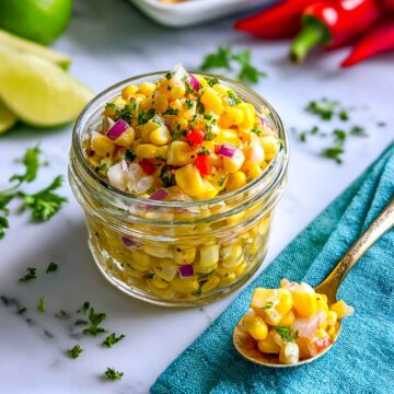 Close-up of fresh corn relish in a glass jar with a gold spoon resting on a teal napkin, showing sweet corn kernels mixed with red onion, peppers, and chopped herbs.