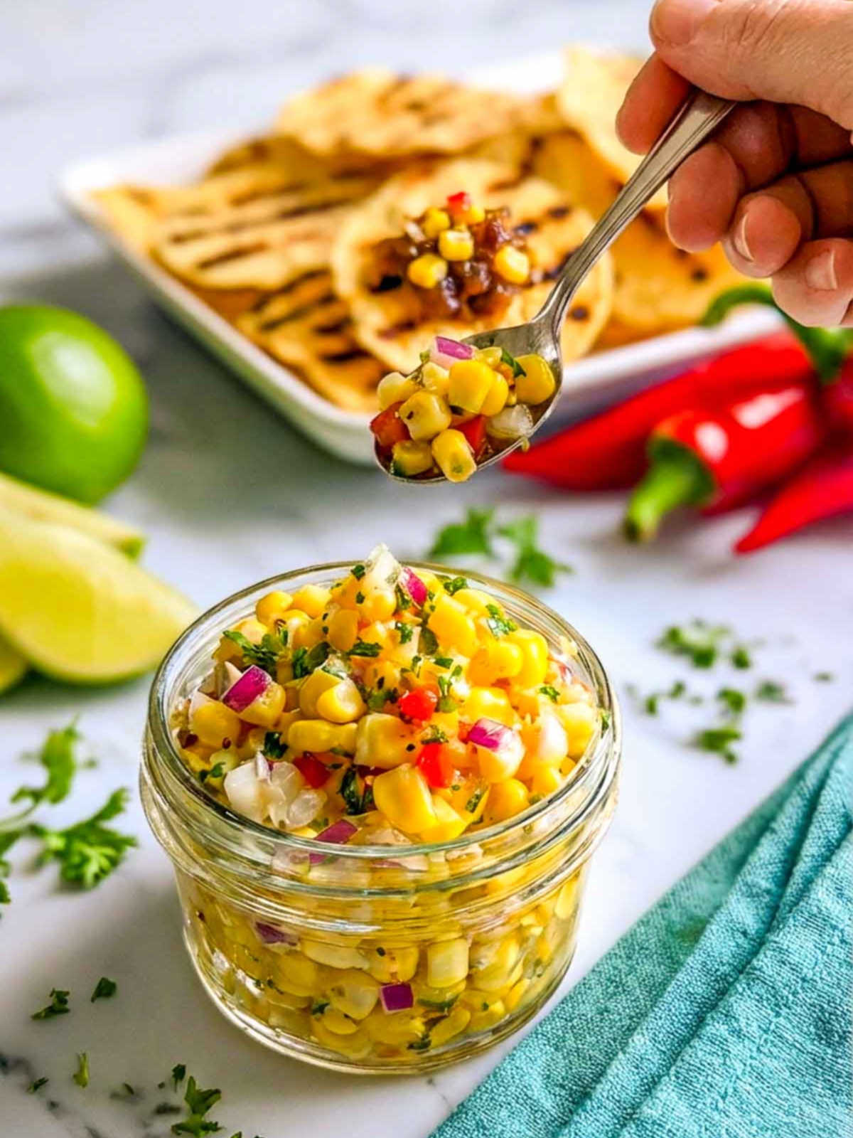 A hand holding a spoonful of vibrant corn relish above a glass jar, with grilled flatbread, red chilies, and lime wedges blurred in the background on a white marble countertop.