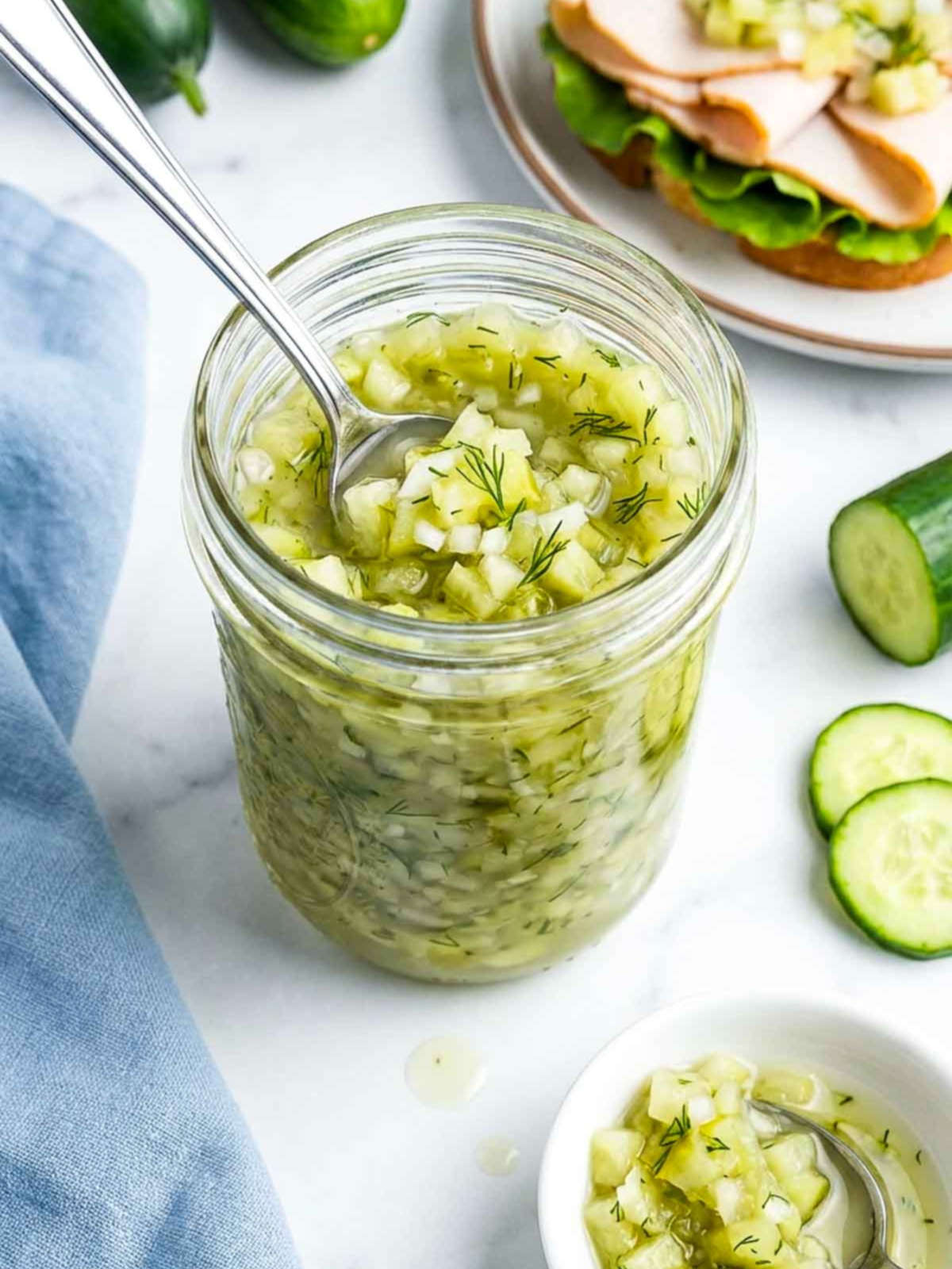 A glass jar filled with homemade cucumber relish sits on a white marble surface with a spoon inside, fresh dill visible throughout, and sliced cucumbers and a turkey sandwich in the background.