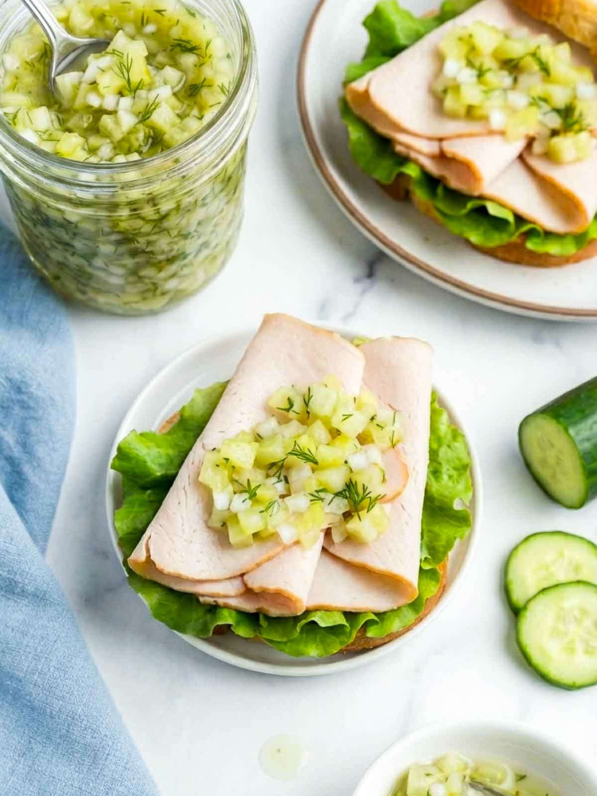 An open-faced turkey sandwich topped with chopped cucumber relish rests on a white plate beside a jar of relish, with cucumber slices and a light blue napkin on a white marble surface.