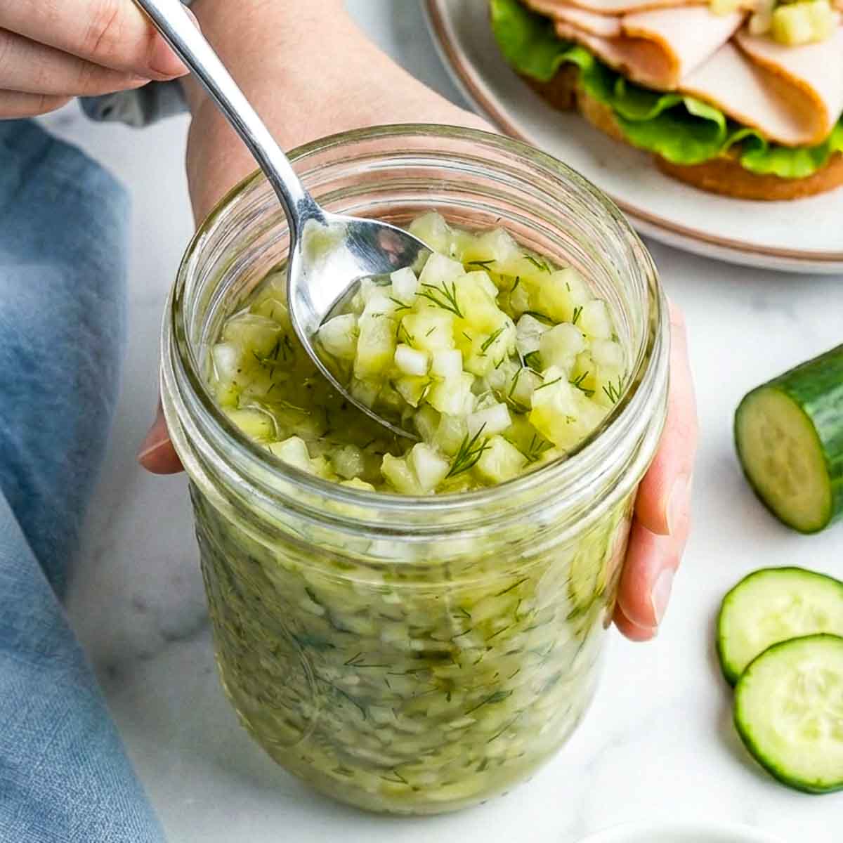 A hand holds a glass jar of cucumber relish while a spoon scoops the chunky mixture, showing finely diced cucumber, onion, and dill on a white marble countertop.