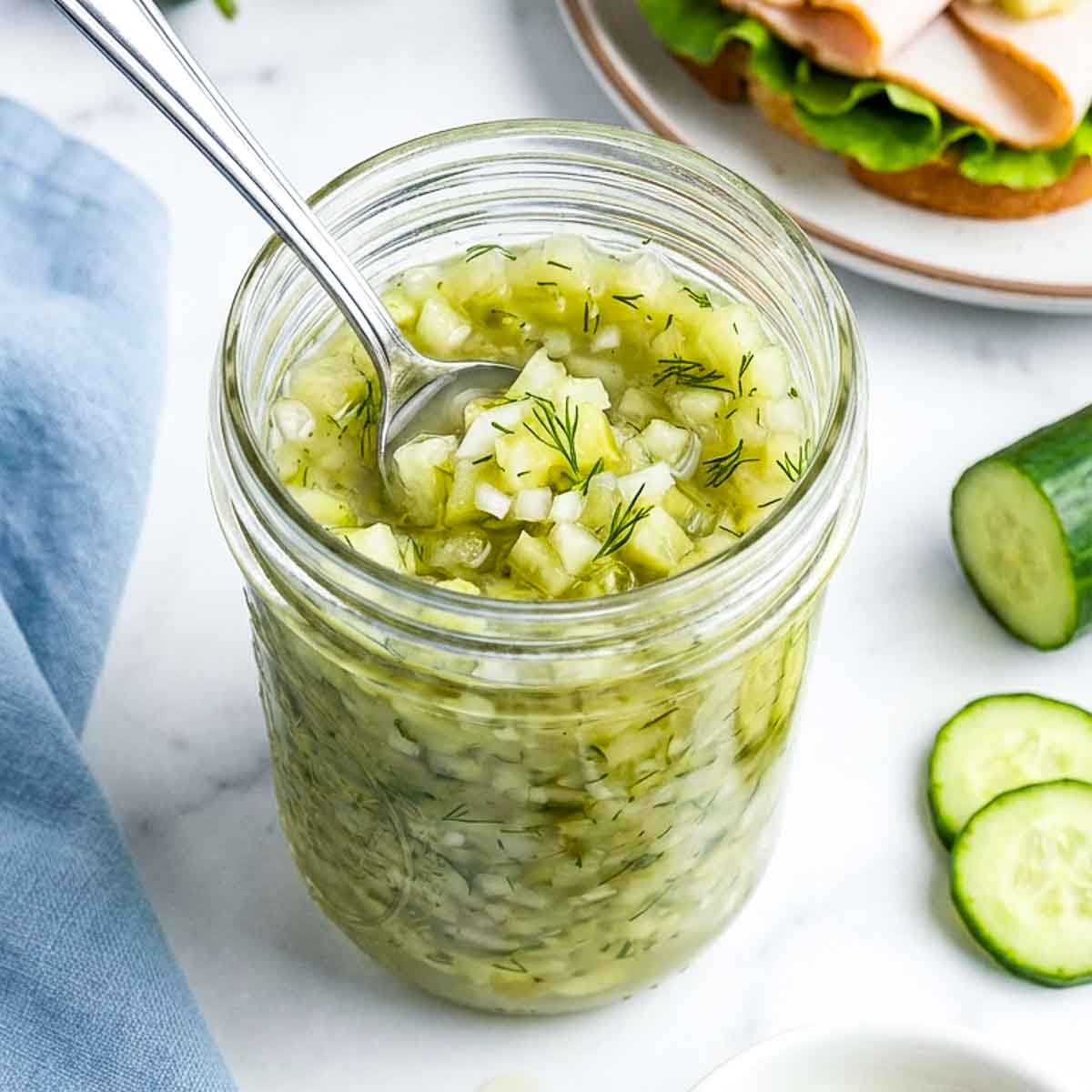 A close-up of a jar of cucumber relish with a spoon resting inside highlights the crisp diced cucumbers and fresh dill, set on a white marble surface with cucumber slices and an open-faced sandwich in the background.
