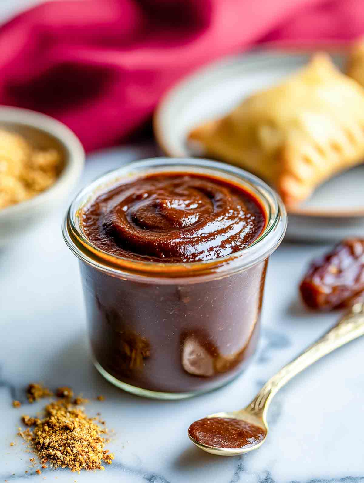A jar of thick date tamarind chutney sits on a marble surface with a spoon, spices, and a samosa in the background.