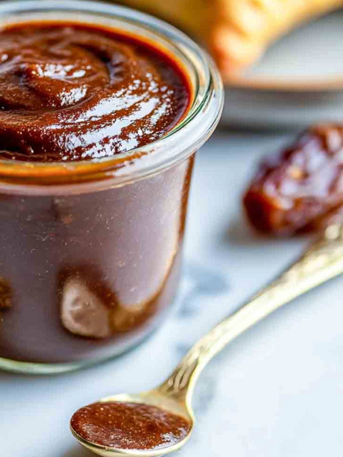 A close-up shows rich date tamarind chutney in a glass jar with a spoonful placed in front on a marble countertop.