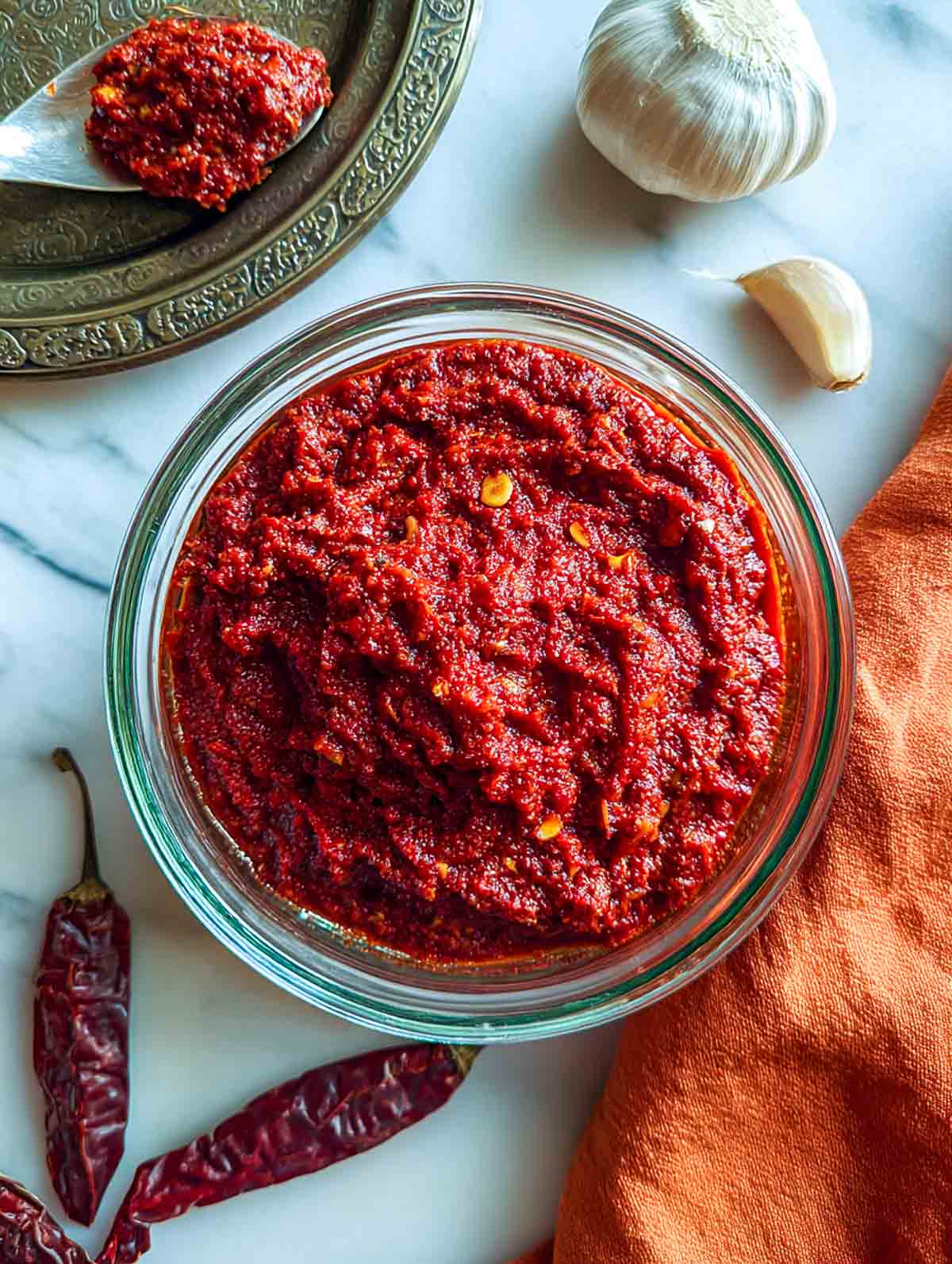 A glass bowl filled with vibrant red garlic chutney sits on a white marble surface, surrounded by dried red chilies, a whole garlic bulb, a loose garlic clove, and an orange cloth napkin.