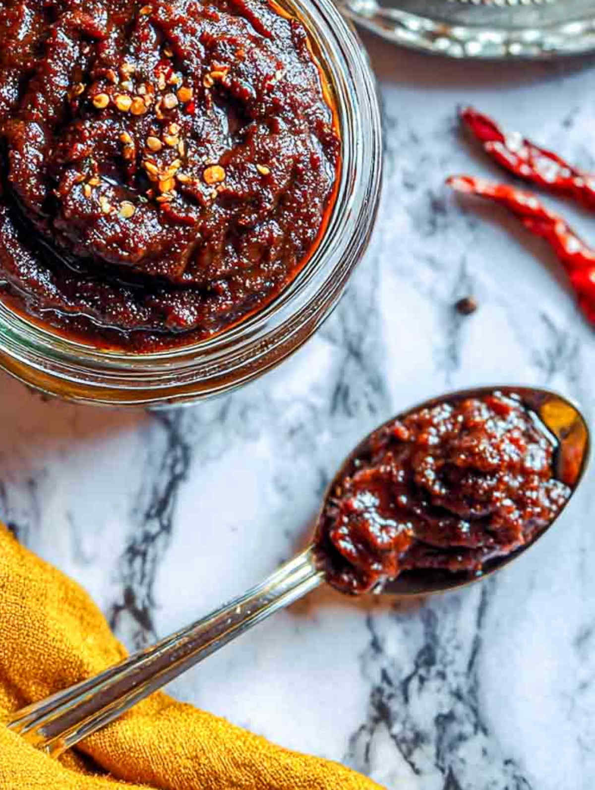 A close-up of a spoonful of rich, glossy ginger chutney on a marble countertop, showing its thick texture and deep reddish-brown color.