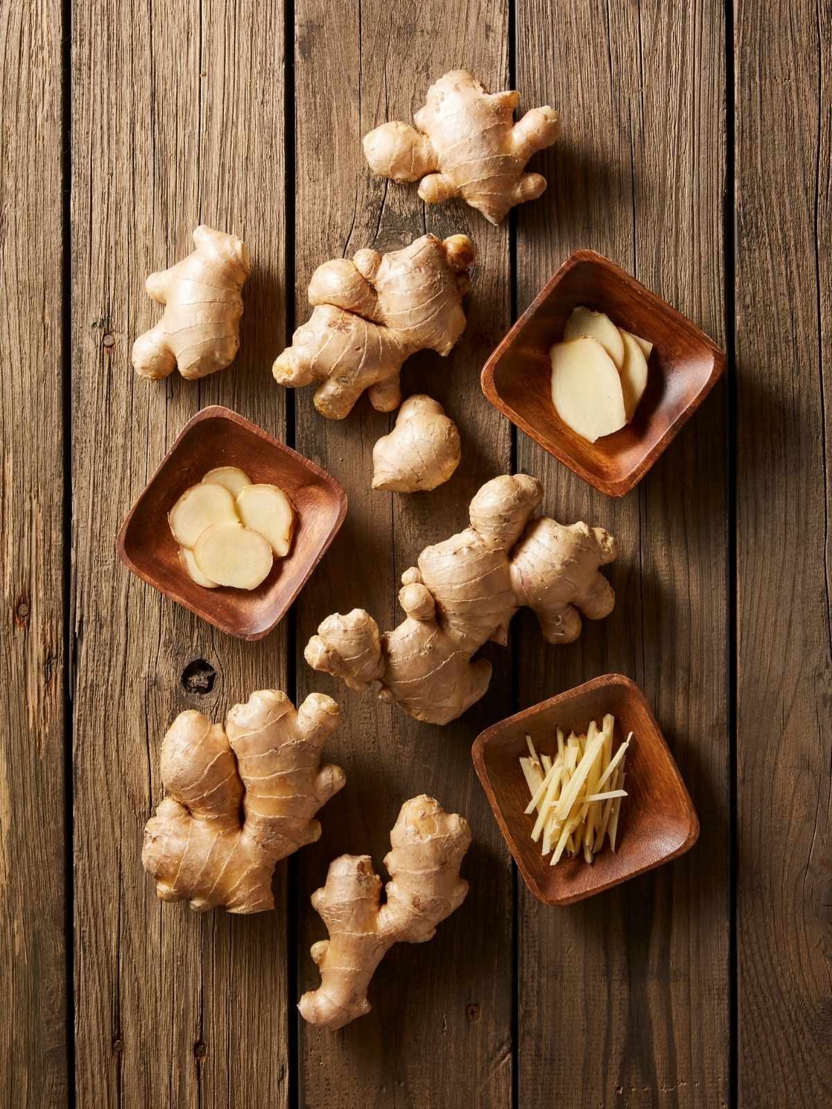 Fresh whole ginger roots with sliced and julienned pieces in small wooden bowls on a rustic wooden surface.