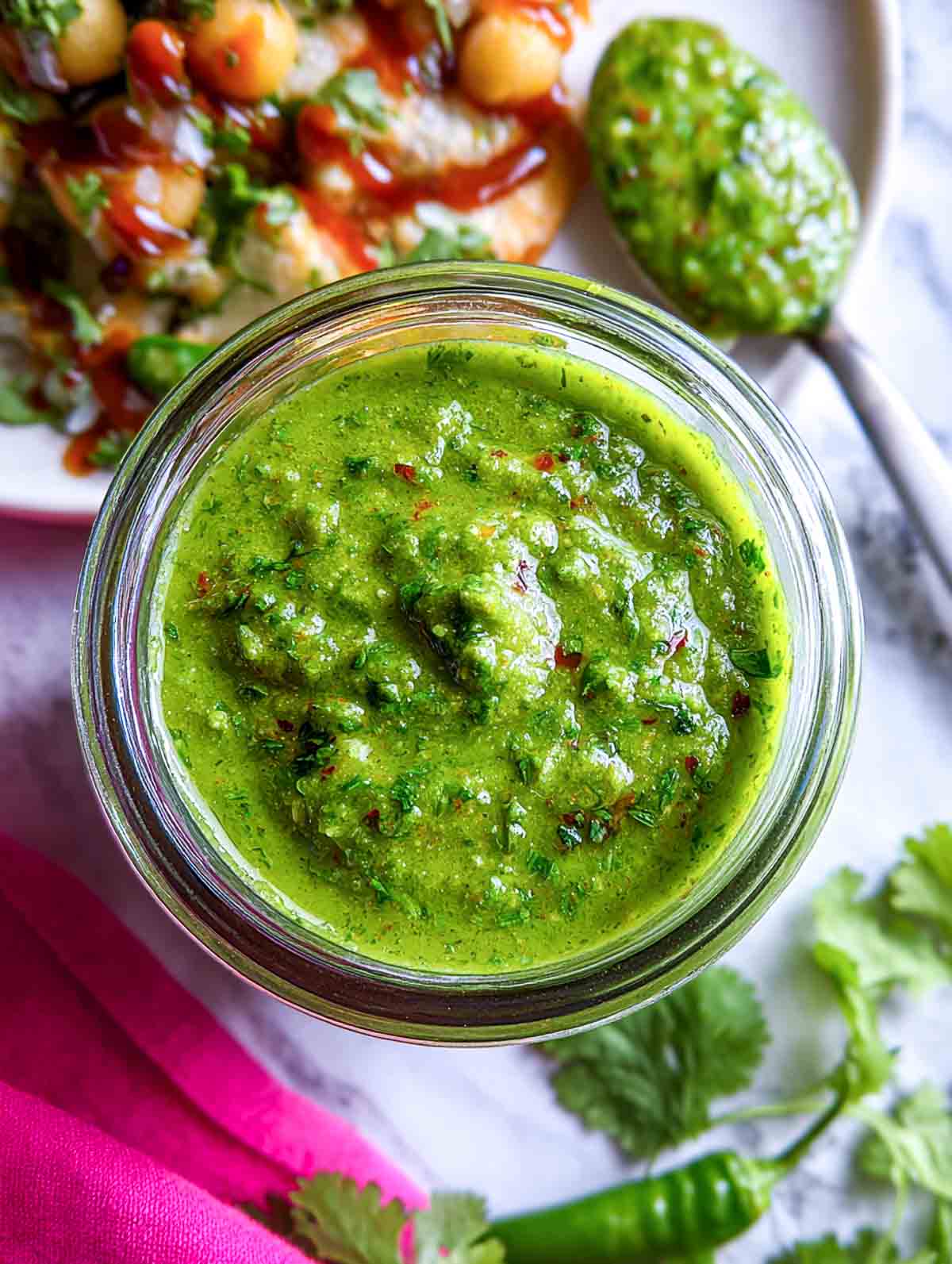 A jar of bright green mint coriander chutney with a thick texture, placed next to fresh cilantro and a pink cloth napkin.