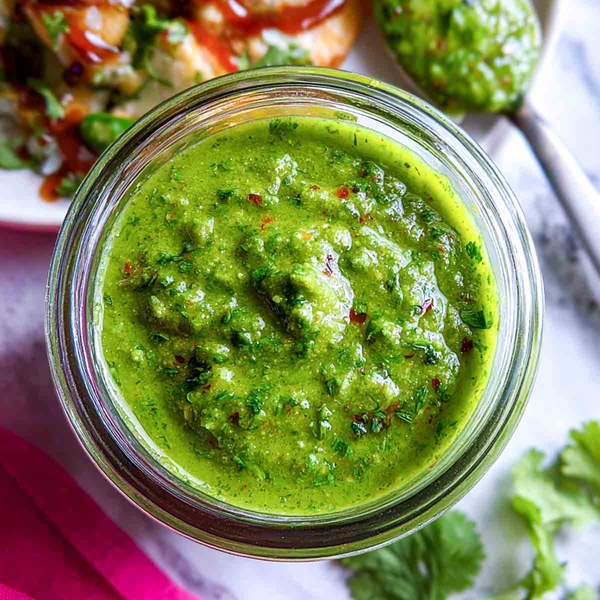 Top view of homemade green chutney in a glass jar with a spoonful served beside chaat topped with chickpeas, yogurt, and tamarind sauce.