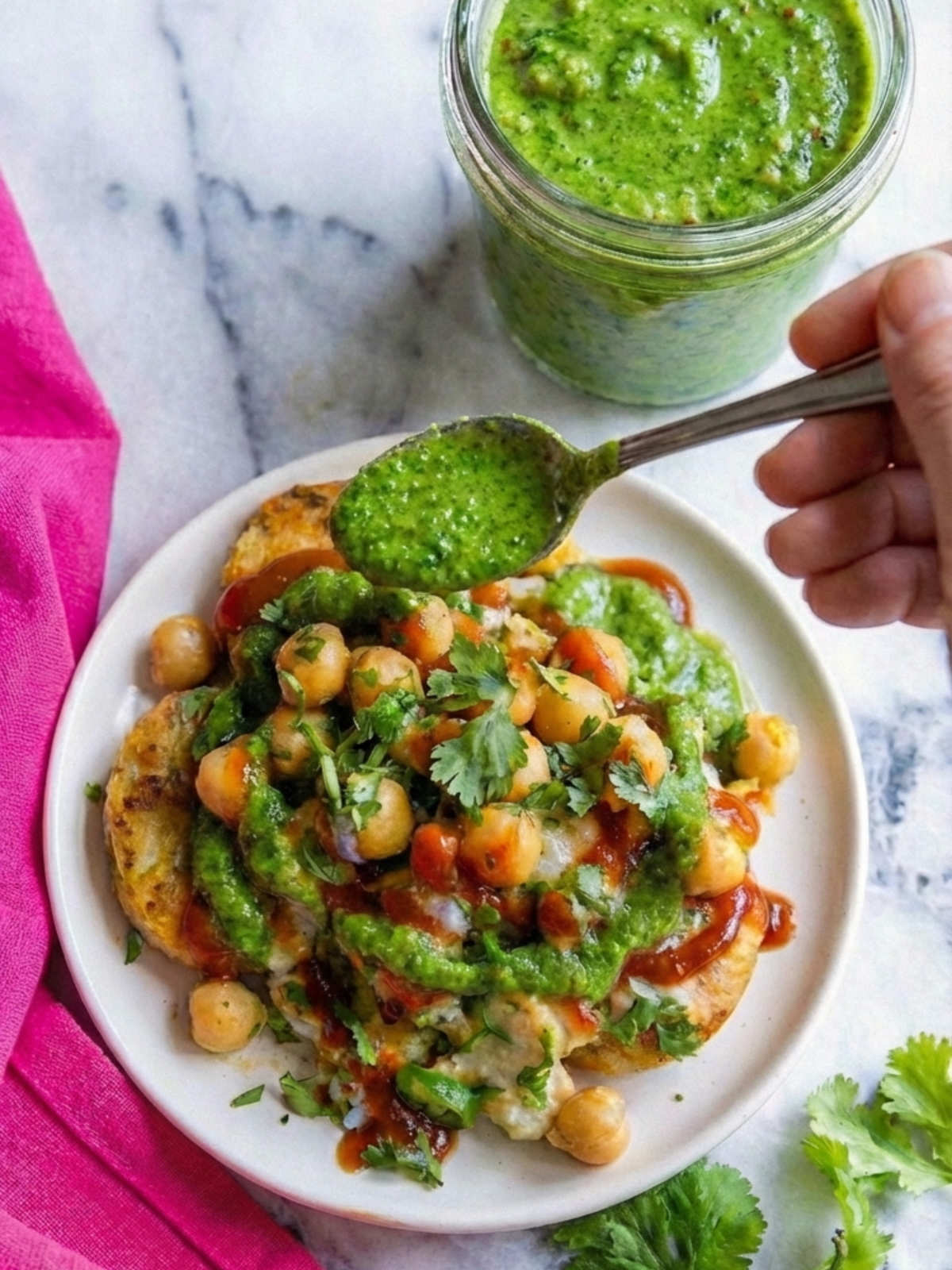 Hand holding a spoonful of green chutney over a plate of chaat layered with chickpeas, yogurt, cilantro, and sweet tamarind sauce.