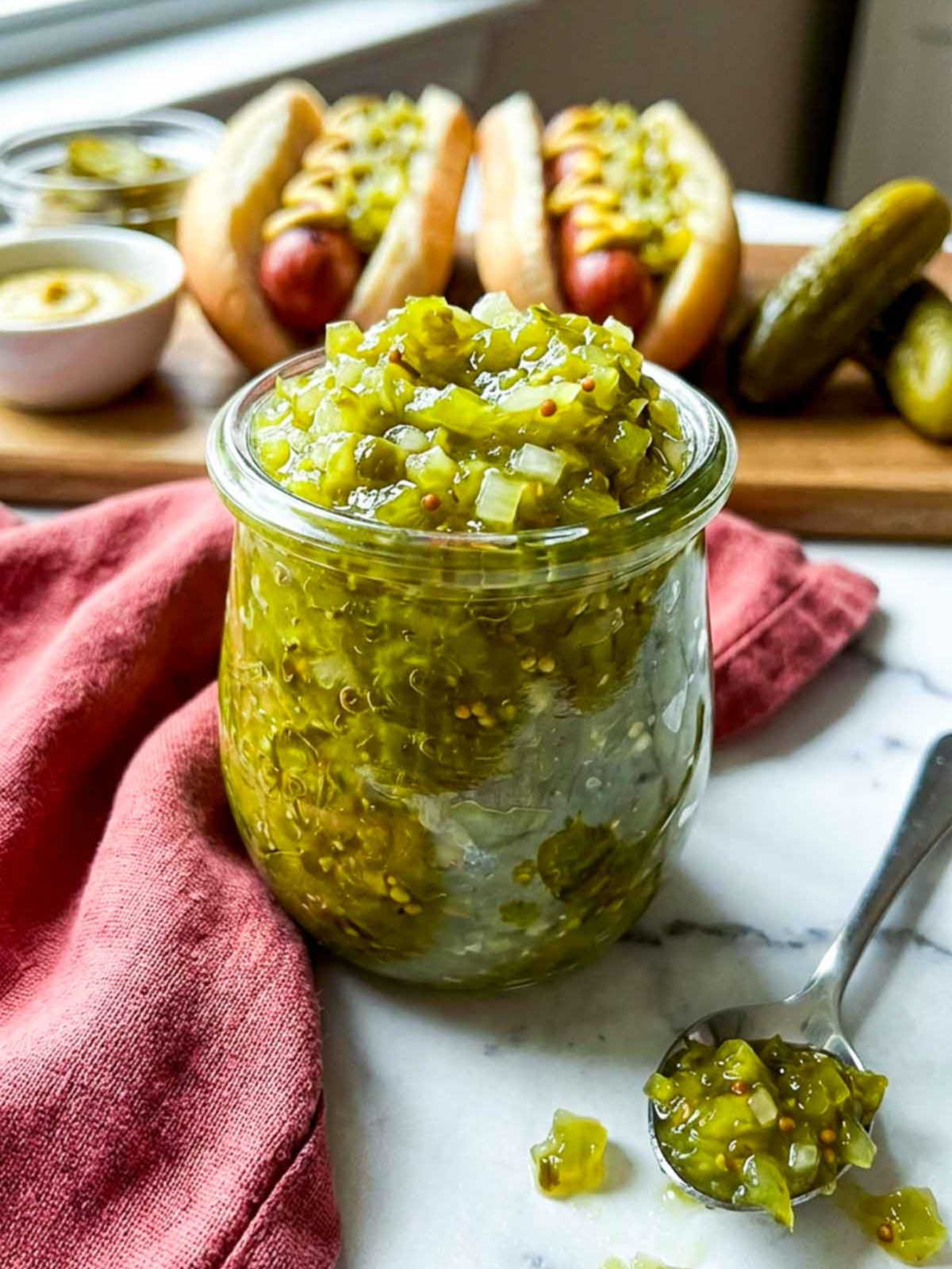 A glass jar filled with chunky homemade hot dog relish sits on a marble countertop with hot dogs and mustard in the background.