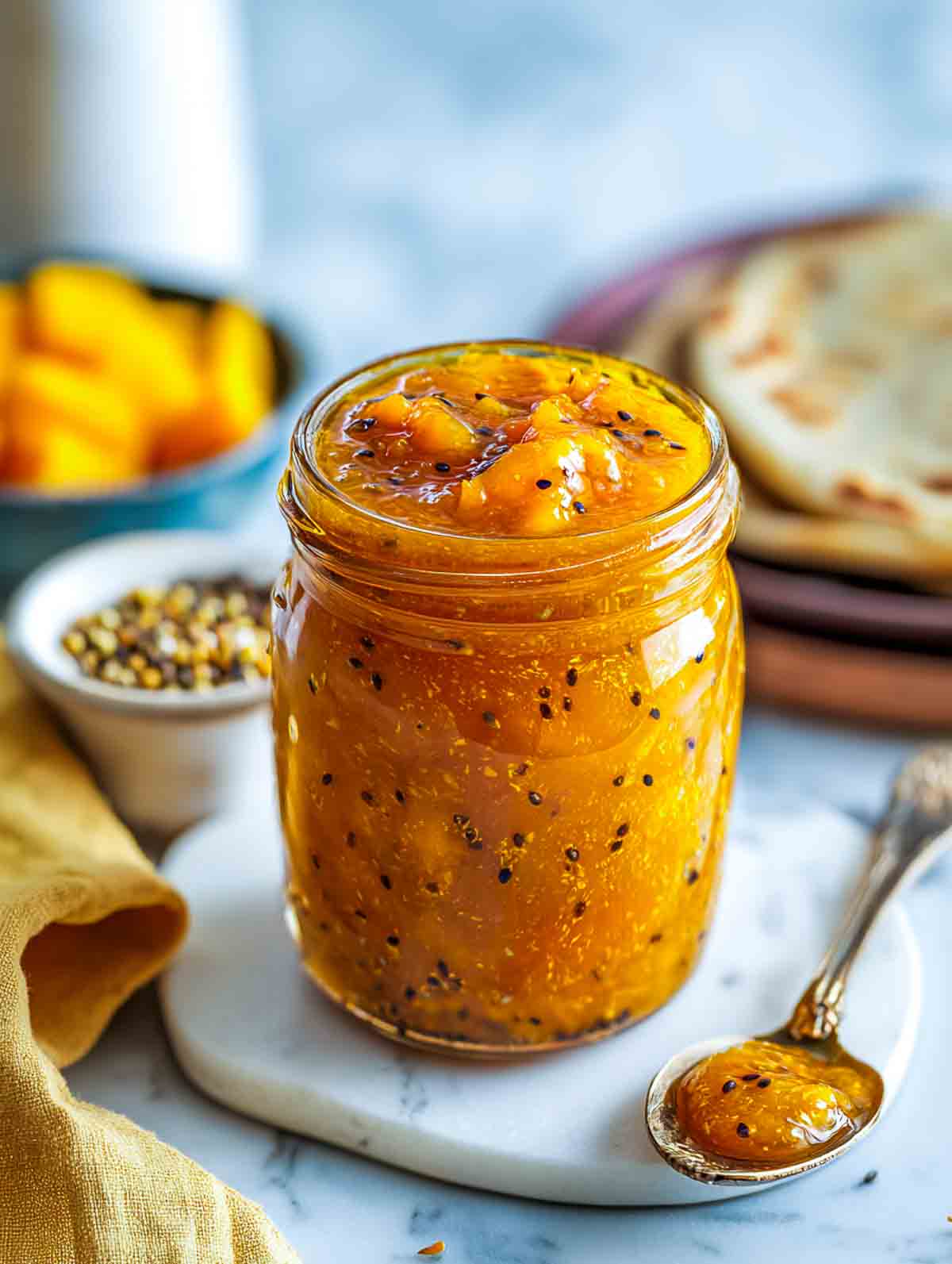 A jar of chunky homemade mango chutney filled with tender mango pieces and black mustard seeds, served on a white marble board with naan bread and whole spices in the background.