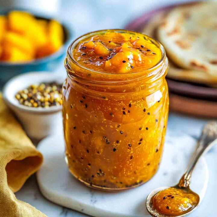A mason jar filled with glossy mango chutney speckled with black mustard seeds, placed on a marble surface with a small bowl of spices and soft naan behind it.