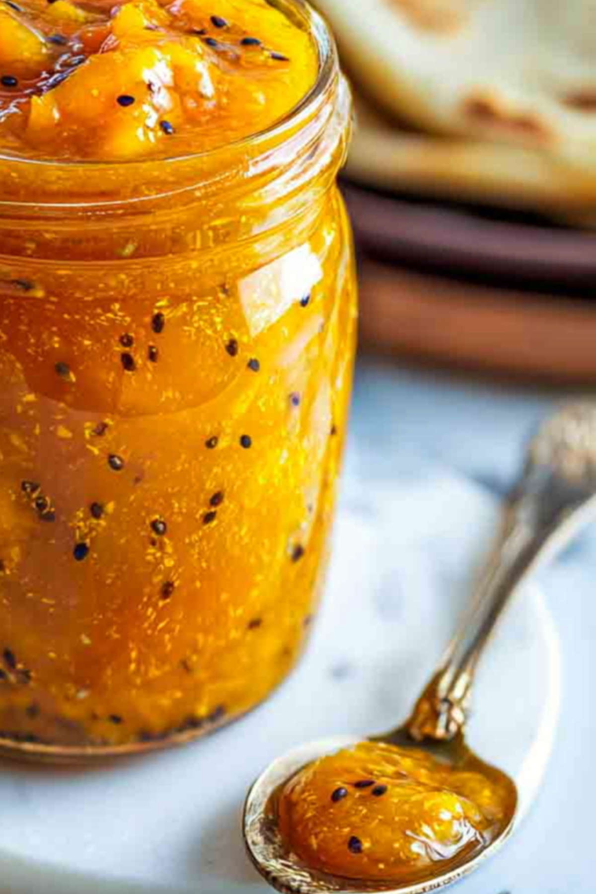 A close-up of thick, golden mango chutney with visible mango chunks and mustard seeds, with a spoonful resting beside the jar on a white marble board.
