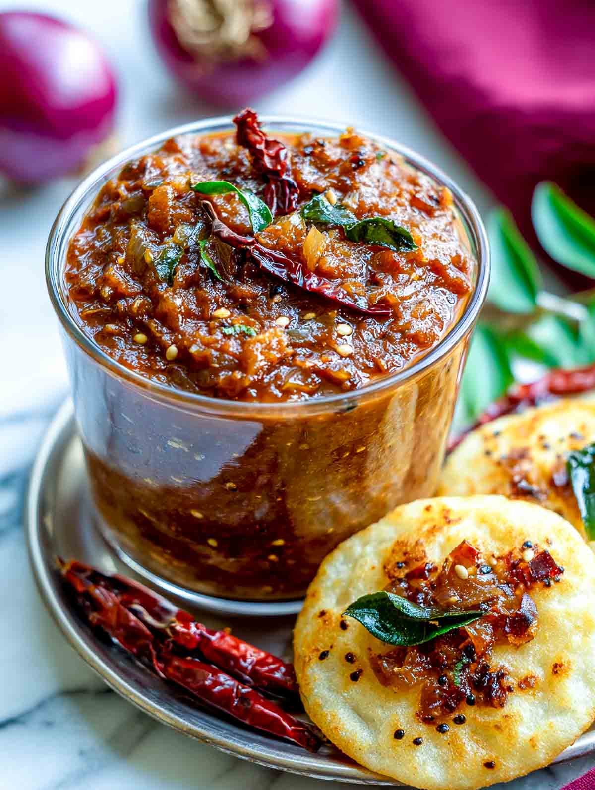 A glass bowl filled with spicy South Indian onion chutney topped with dried red chilies and curry leaves, served with mini idlis on a steel plate.