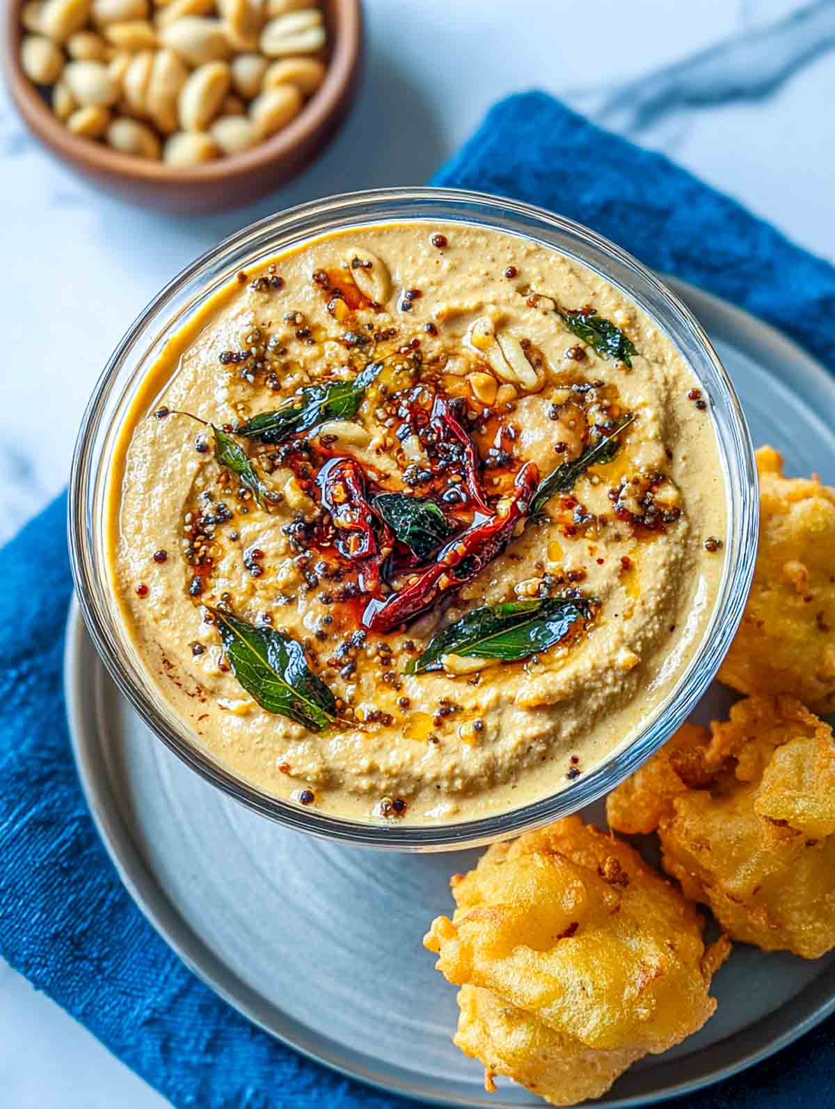 A bowl of creamy peanut chutney topped with tempered mustard seeds, dried red chilies, curry leaves, and garlic, served with golden fritters on a plate.