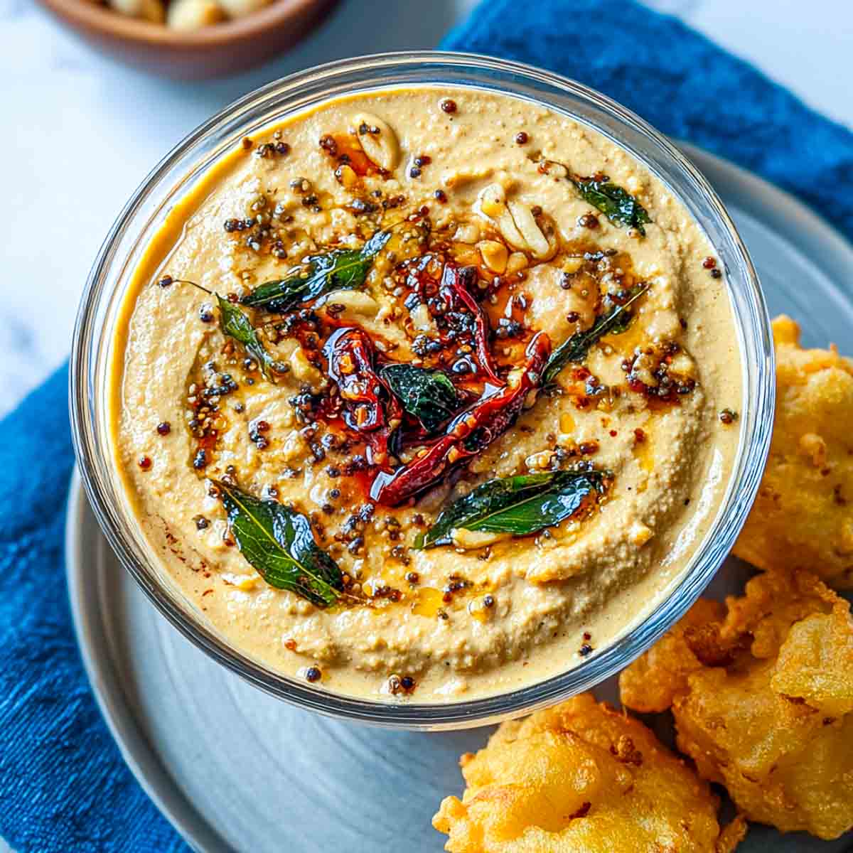 Close-up of smooth peanut chutney with a spiced oil tempering of mustard seeds, curry leaves, garlic, and red chilies in a glass bowl.