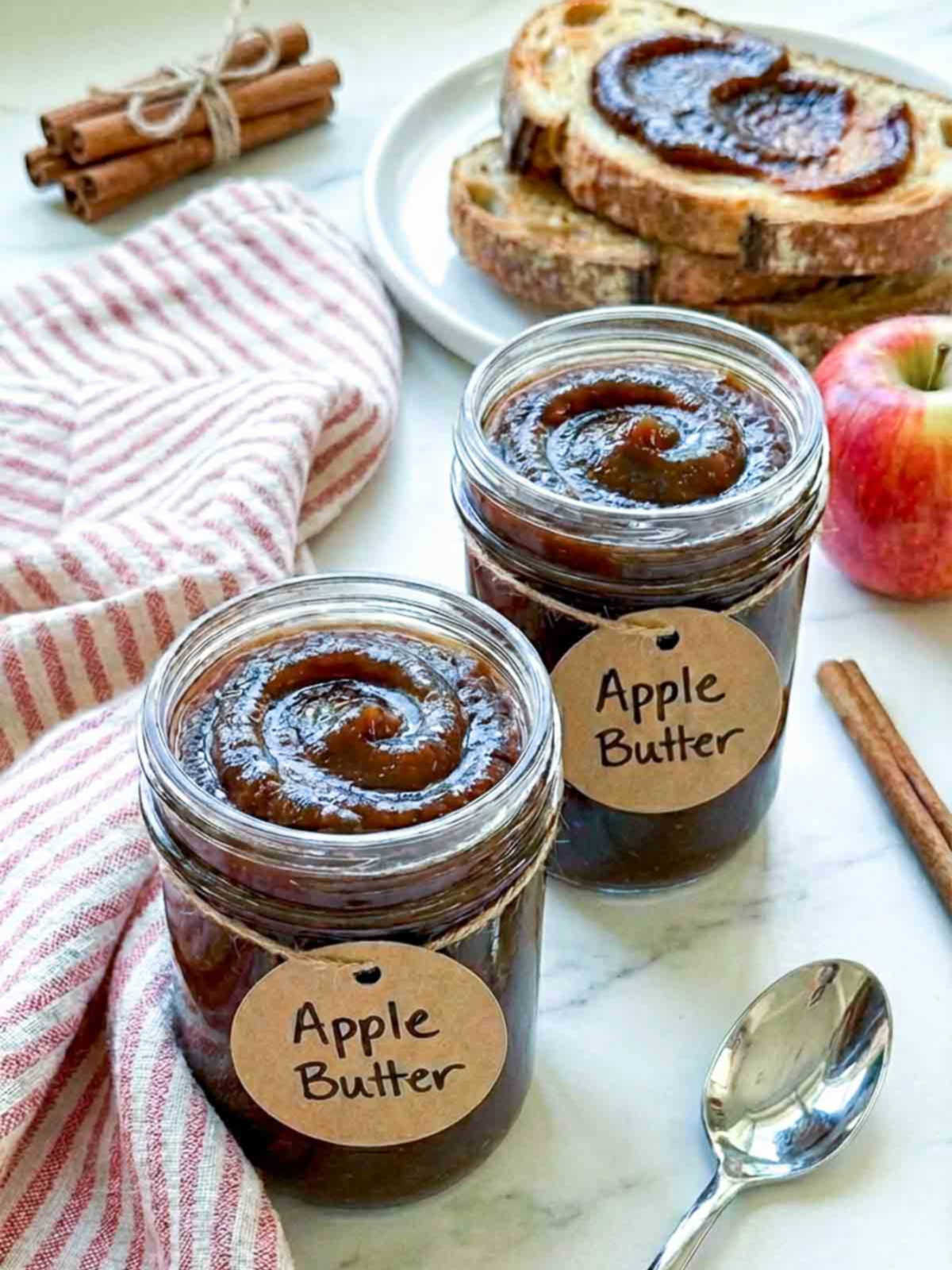 Two jars of apple butter with labeled tags, served with toast, apples, and cinnamon sticks.