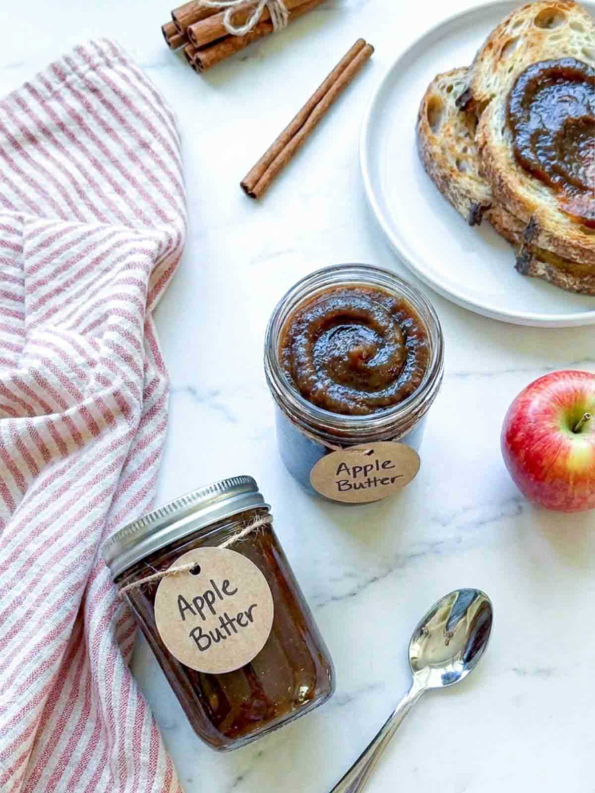 Jar of apple butter with labeled tag, served with toast, apple, and cinnamon sticks.
