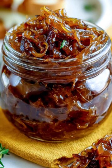 Close-up of a jar filled with caramelized onion relish on a yellow cloth with a spoon beside it.