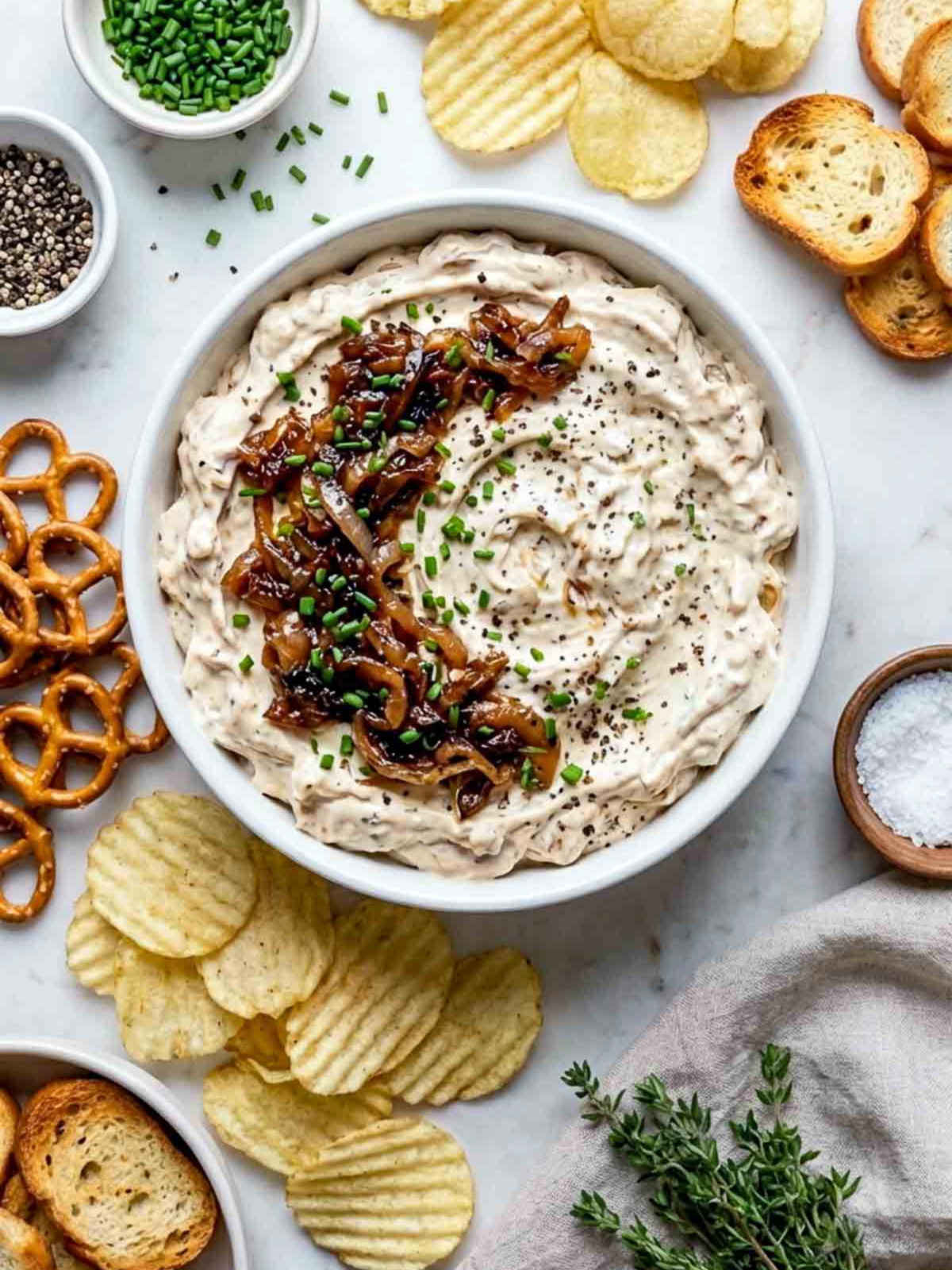 Creamy caramelized onion dip topped with sautéed onions and chives, surrounded by chips, pretzels, and toasted bread.