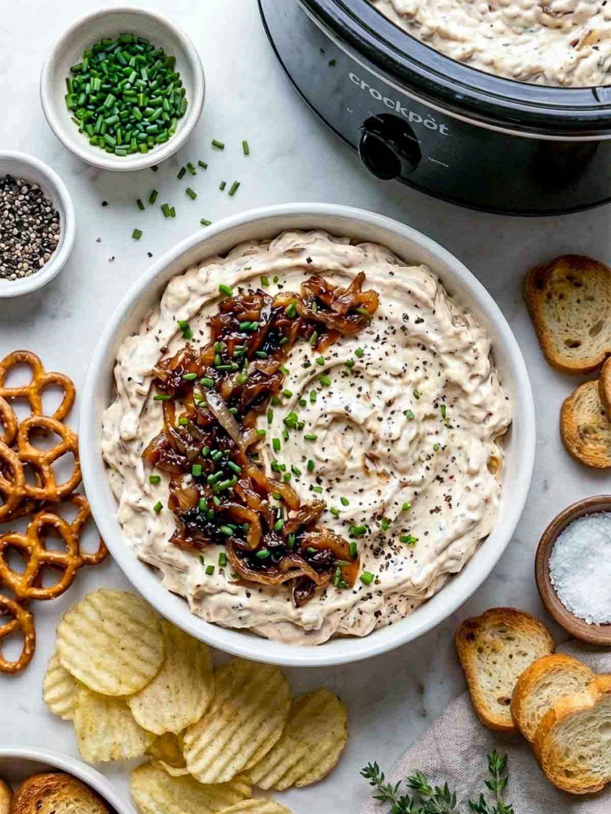 Creamy caramelized onion dip topped with sautéed onions and chives, served with chips, pretzels, and toasted bread beside a crockpot.