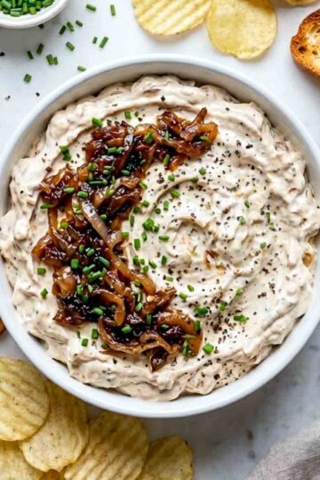 Top view of creamy caramelized onion dip garnished with sautéed onions, chives, and black pepper, surrounded by chips, pretzels, and toasted bread.