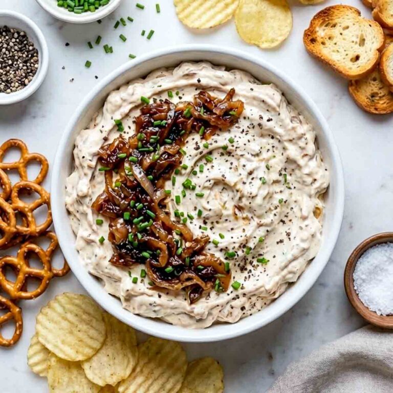 Top view of creamy caramelized onion dip garnished with sautéed onions, chives, and black pepper, surrounded by chips, pretzels, and toasted bread.