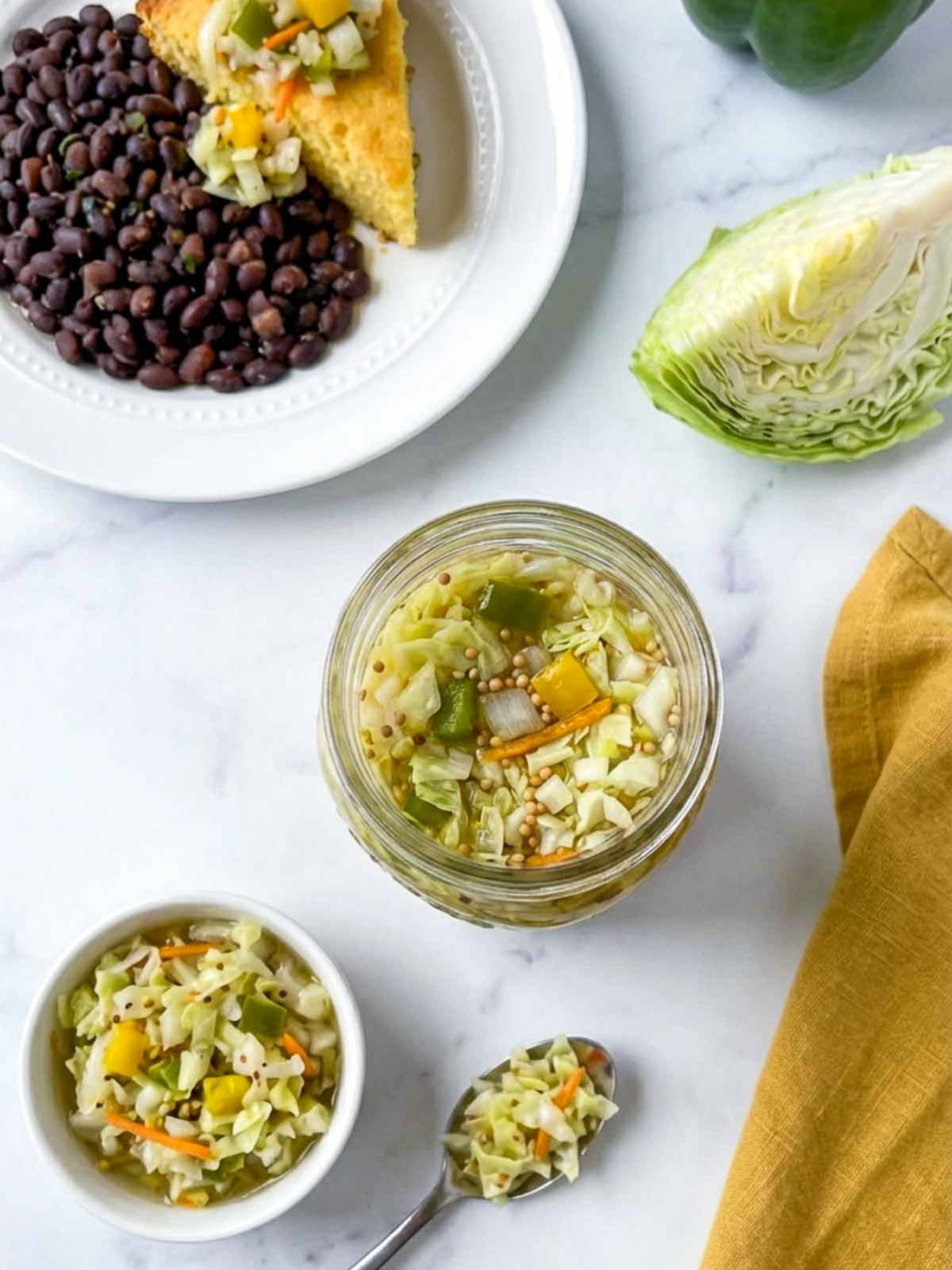 Jar and bowl of chow chow relish with cabbage, peppers, and carrots beside beans and cornbread.