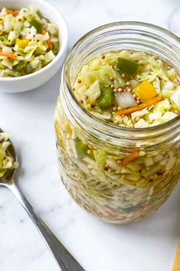 Jar of chow chow relish with cabbage, peppers, and carrots beside a spoon and small bowl.
