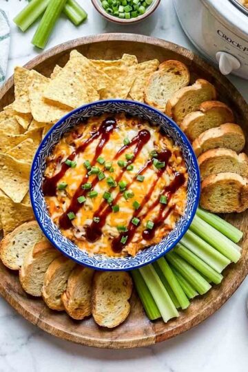 Crockpot BBQ chicken dip with cheese and BBQ sauce, topped with green onions, surrounded by tortilla chips, toasted bread, celery, and shredded cheese, with a bowl of BBQ sauce on the side.