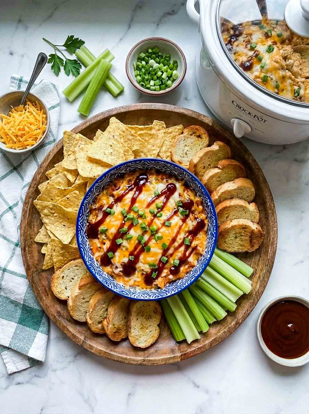 Crockpot BBQ chicken dip served with tortilla chips, toasted bread, celery, green onions, and shredded cheese, with a bowl of barbecue sauce on the side.