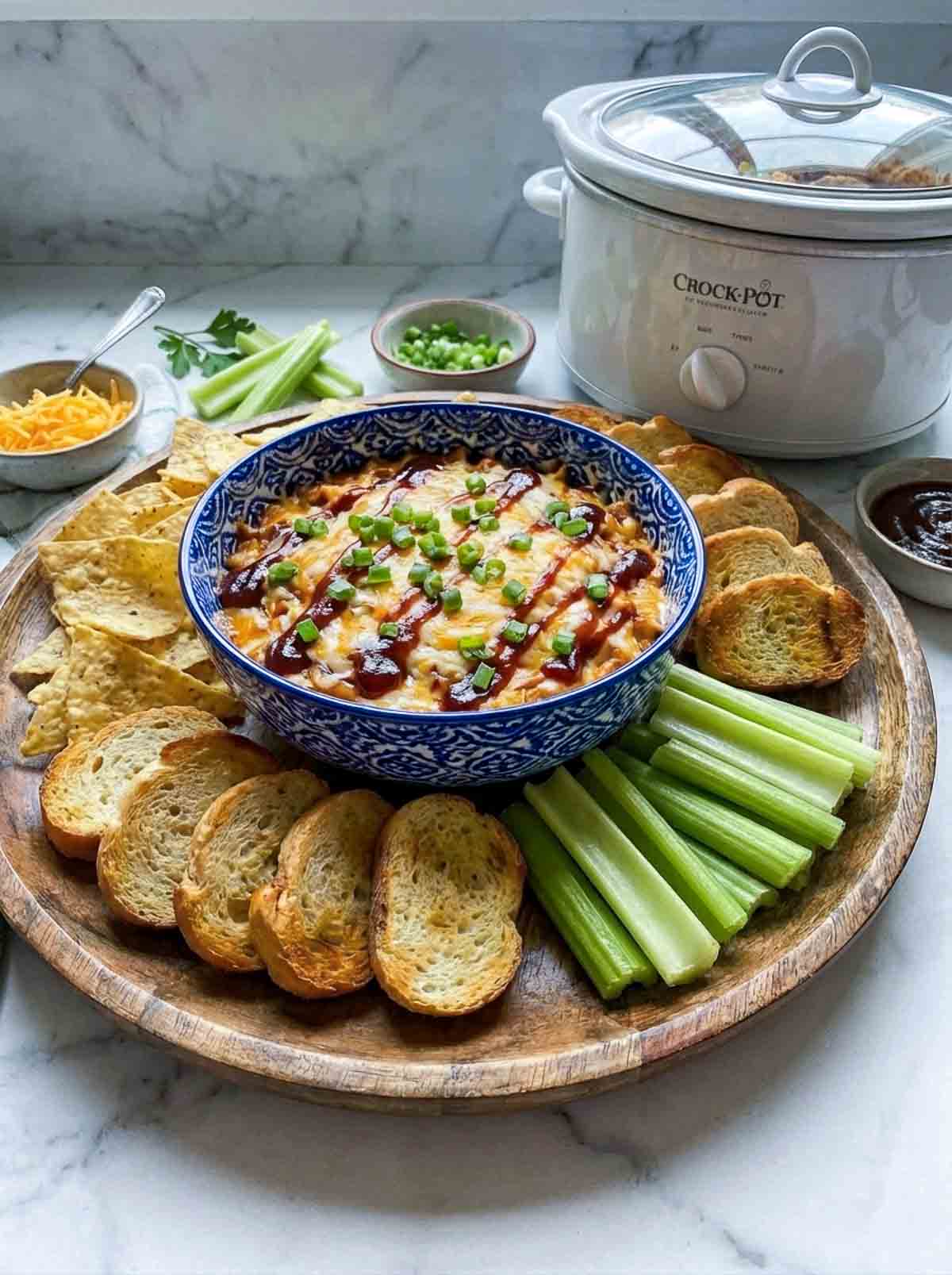 Crockpot BBQ chicken dip garnished with green onions and BBQ sauce, served with tortilla chips, toasted bread, celery, and shredded cheese, with a bowl of BBQ sauce on the side.