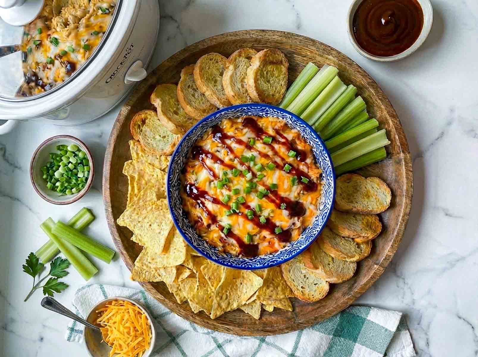 Crockpot BBQ chicken dip with cheese and BBQ sauce, topped with green onions, served with tortilla chips, toasted bread, celery, and shredded cheese on the side, with a bowl of BBQ sauce.