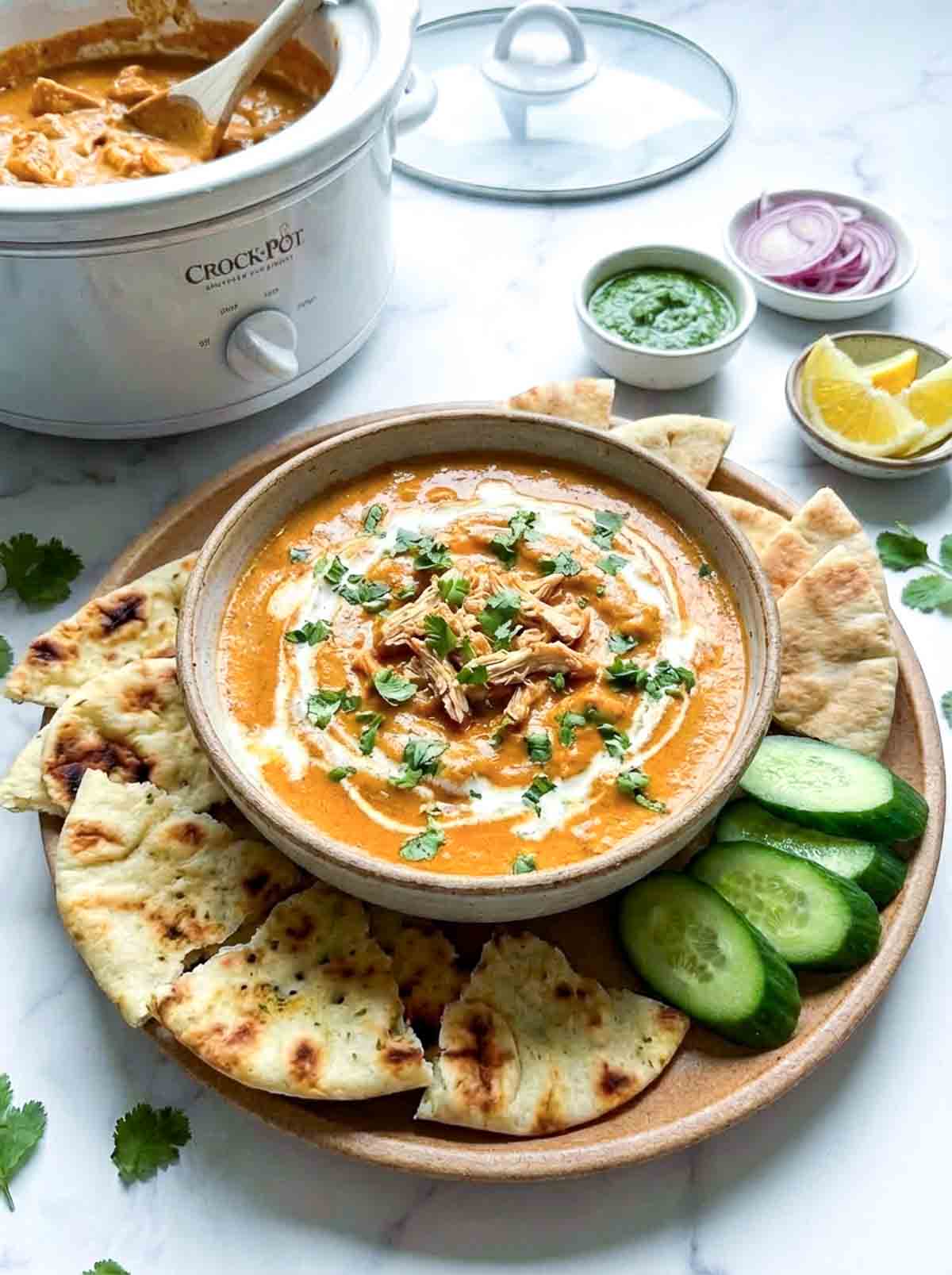 Crockpot butter chicken dip topped with cilantro and cream, served with naan bread, cucumber slices, green chutney, sliced onions, and lemon wedges.