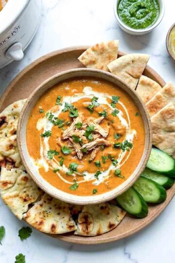Crockpot butter chicken dip with cilantro and cream, topped with shredded chicken, served with naan bread, cucumber slices, green chutney, sliced onions, and lemon wedges.