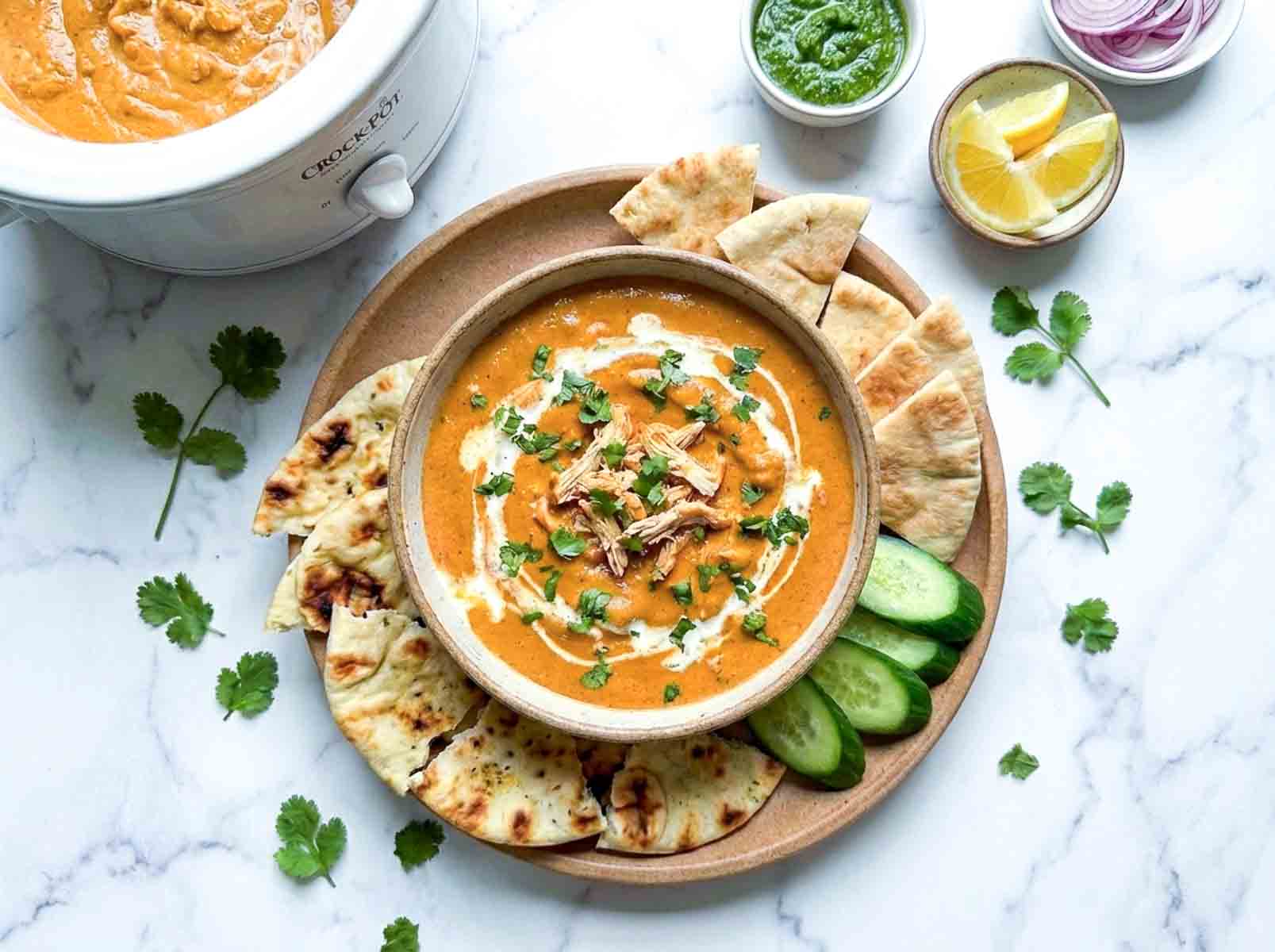Crockpot butter chicken dip with cilantro and cream, served with naan bread, cucumber slices, green chutney, sliced onions, and lemon wedges.