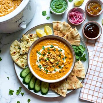 Crockpot butter paneer dip topped with crumbled paneer and cilantro, served with naan bread, cucumber slices, green chutney, pickled onions, mango chutney, and lemon wedges.