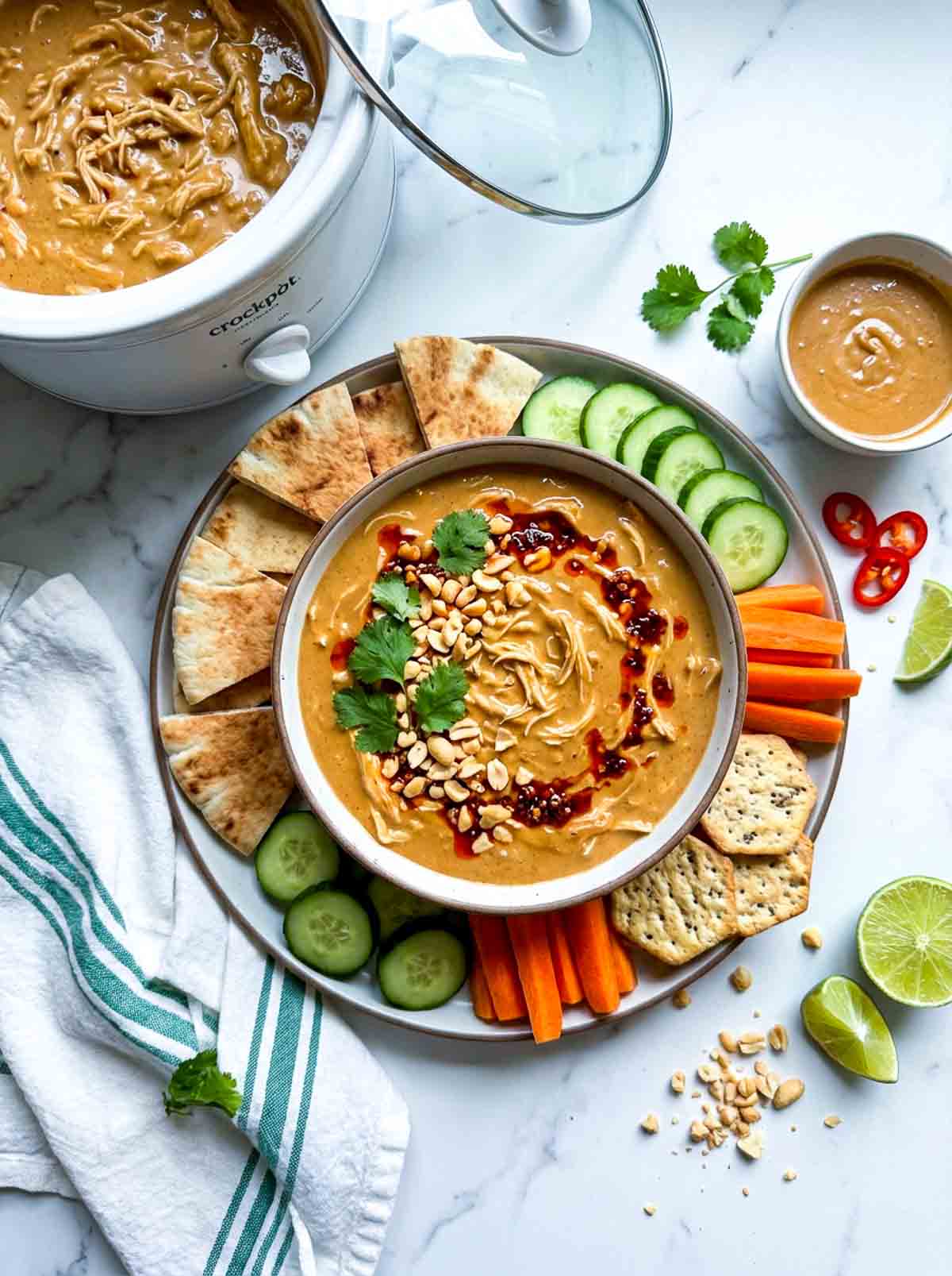 Crockpot peanut chicken dip topped with cilantro, peanuts, and chili sauce, served with pita bread, cucumber slices, carrot sticks, crackers, lime wedges, and a side of peanut sauce.