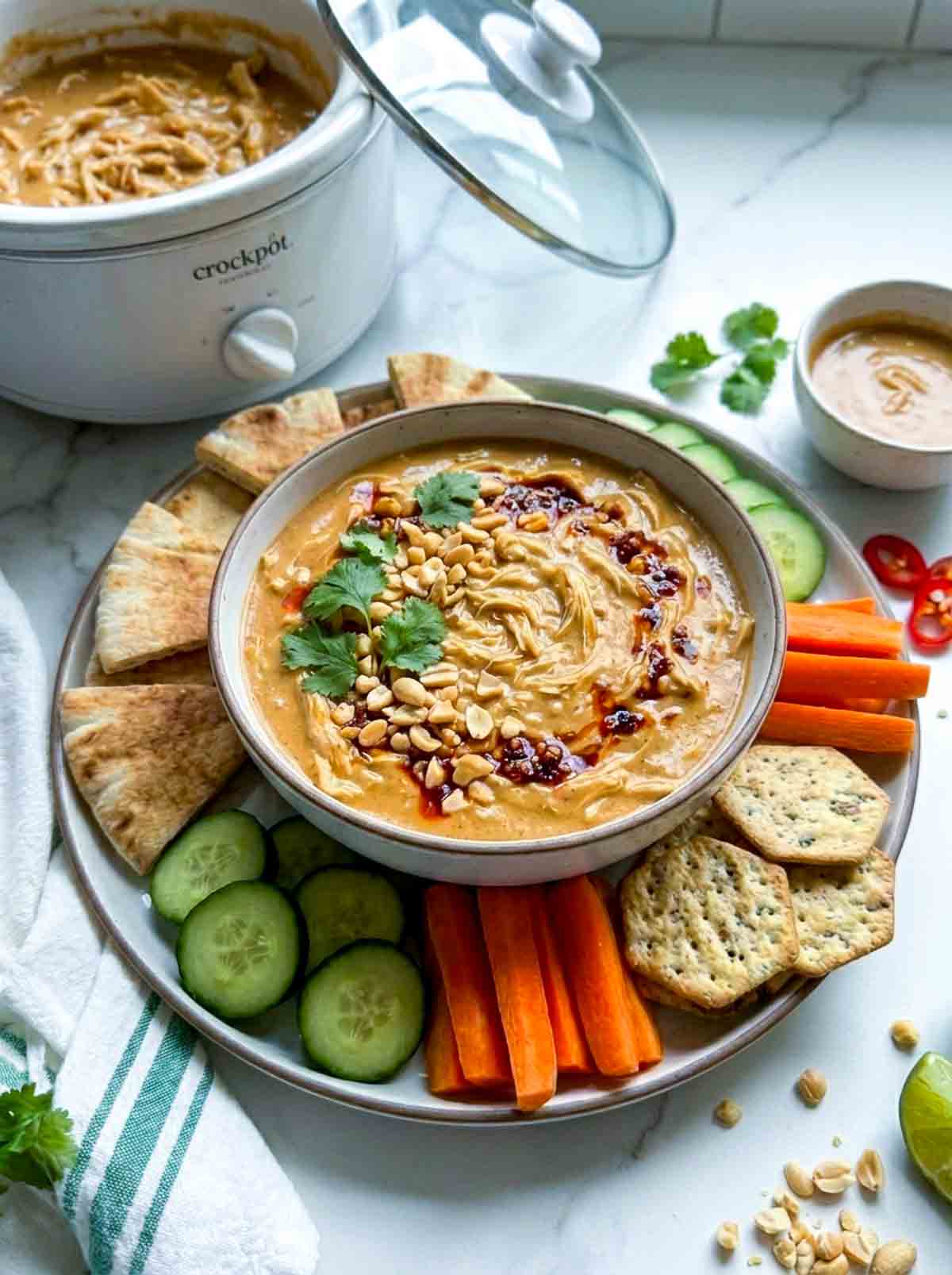 Plate of Crockpot Peanut Chicken Dip served with pita bread, cucumber slices, carrot sticks, and crackers. The dip is garnished with peanuts, cilantro, and a drizzle of sauce, with additional ingredients like lime wedges, sliced chili peppers, and dipping sauces placed around the plate. In the background, a slow cooker filled with the dip is visible