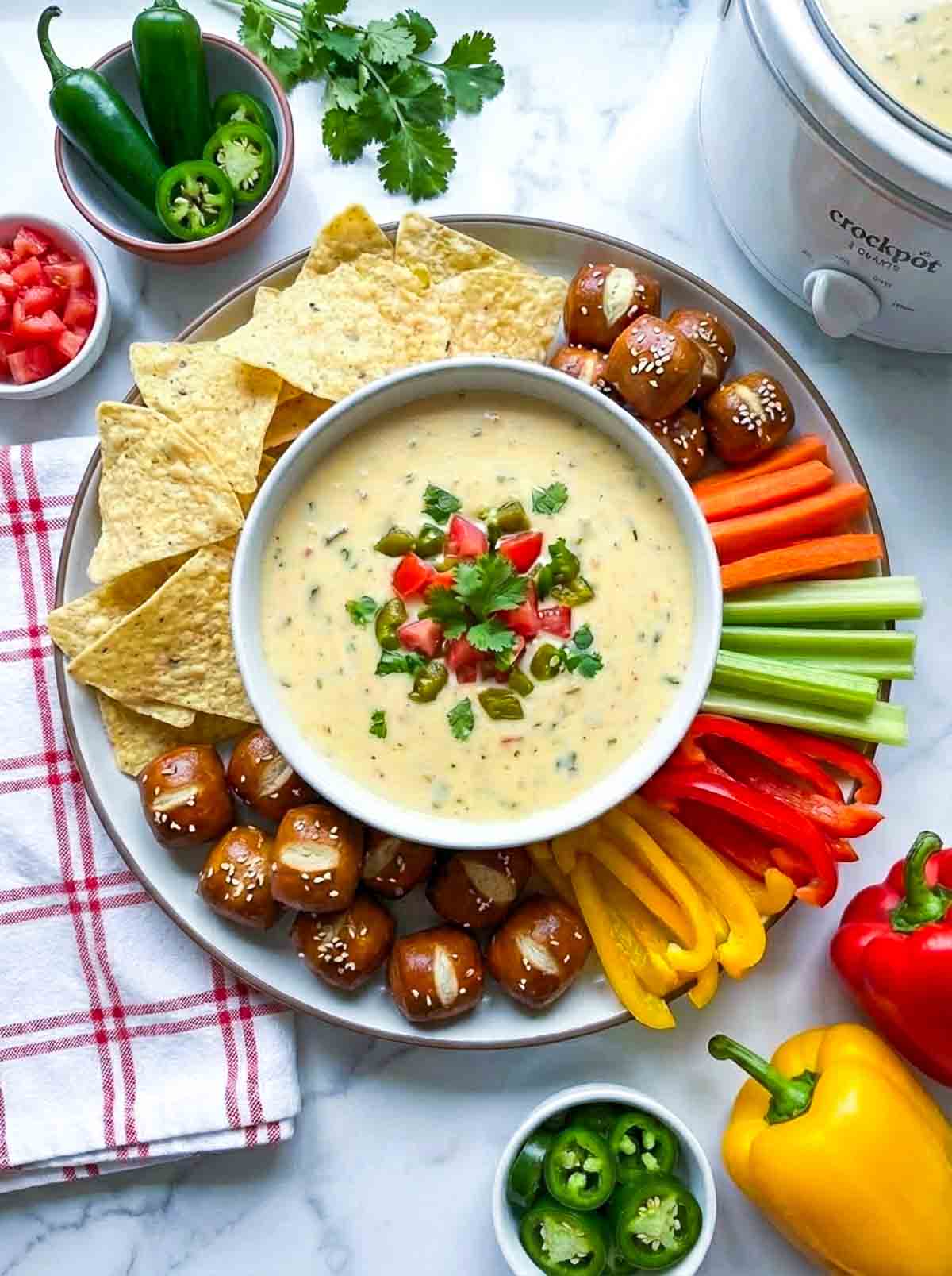 Plate of crockpot queso dip surrounded by tortilla chips, pretzel bites, and sliced veggies like carrots, celery, and bell peppers, with bowls of sliced jalapeños and diced tomatoes.
