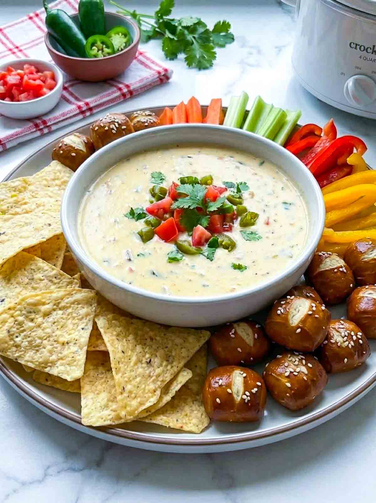 Plate of crockpot queso dip topped with cilantro, tomatoes, and jalapeños, surrounded by tortilla chips, pretzel bites, and sliced veggies like carrots, celery, and bell peppers.