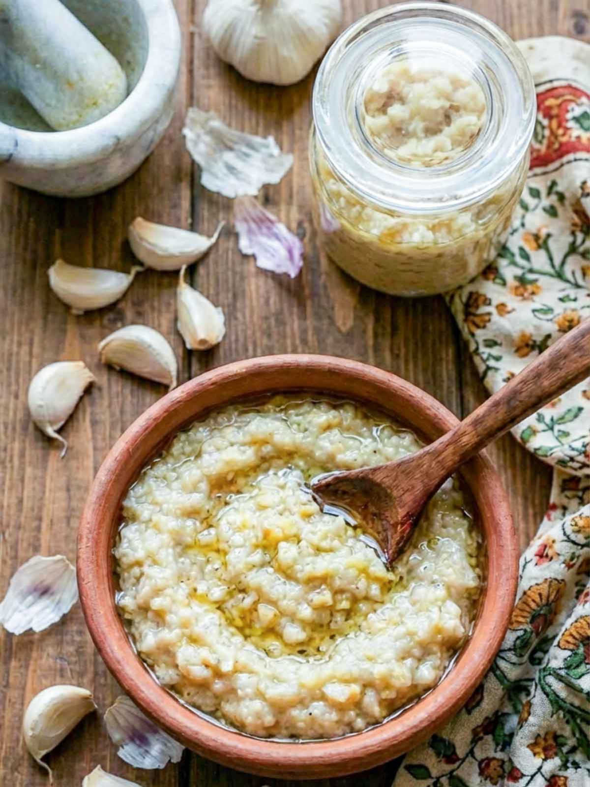 Bowl of garlic paste with olive oil, surrounded by fresh garlic cloves, a mortar and pestle, and a jar of minced garlic on a wooden table.